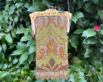 May include: Vintage metal mesh purse with a decorative floral pattern in shades of yellow, orange, and brown. The purse has a silver chain handle and a decorative frame with orange and green accents. The purse is hanging in front of a green leafy background.