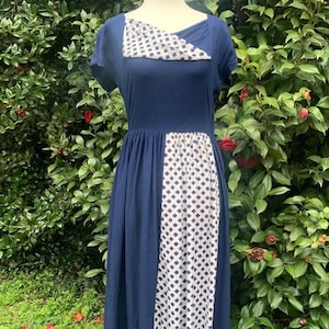 May include: A navy blue dress with a white and navy blue patterned panel on the front. The dress has short sleeves and a unique neckline with a patterned accent. The dress is displayed on a mannequin.