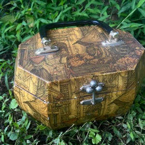 Op de afbeelding: Vintage achthoekige doos met een zwarte handgreep en zilverkleurige hardware. De doos heeft een gedetailleerde illustratie in bruin- en beigetinten, die een historische scène uitbeeldt. De doos ligt op groen gebladerte.