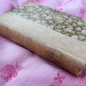 May include: An antique book with a floral patterned cover and spine. The cover features a design of white flowers and green leaves on a brown background. The spine is a light tan color with gold embossed floral detailing and the number "146".