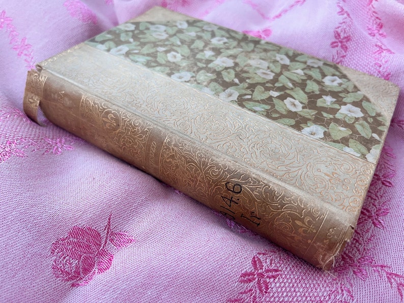 May include: An antique book with a floral patterned cover and spine. The cover features a design of white flowers and green leaves on a brown background. The spine is a light tan color with gold embossed floral detailing and the number "146".