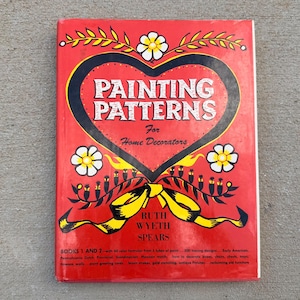 以下が含まれることがあります： Ruth Wyeth Spears著の赤いハードカバー本「Painting Patterns For Home Decorators」。表紙には大きな黒いハートの輪郭、黄色い花のモチーフ、黄色いリボンのグラフィックが描かれています。テキストには「Books 1 and 2」の詳細が含まれています。