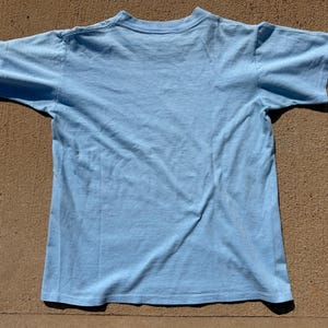 以下が含まれることがあります： クルーネックの半袖ライトブルーTシャツ。衣服は平らに置かれており、シャツの背面が見えています。衣服の表面全体が単色のライトブルーの生地で構成されています。