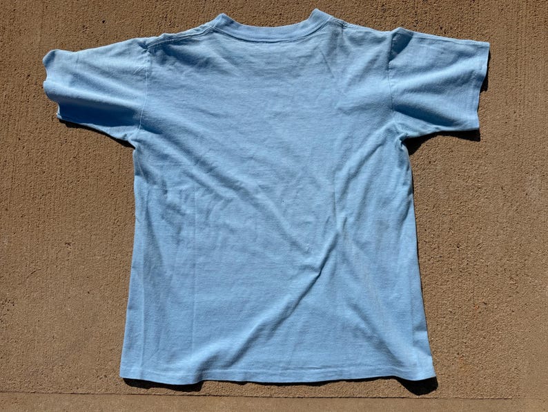 以下が含まれることがあります： クルーネックの半袖ライトブルーTシャツ。衣服は平らに置かれており、シャツの背面が見えています。衣服の表面全体が単色のライトブルーの生地で構成されています。