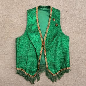 May include: A green vest with gold trim and fringe. The vest has a textured fabric and is decorated with colourful accents along the edges. Two gold-coloured buttons are on the upper right side.