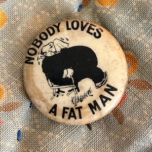 May include: A circular off-white pinback button featuring a black ink illustration of a person sitting in a chair and crying. The text NOBODY LOVES A FAT MAN is printed in black capital letters around the graphic, with a signature at the bottom.