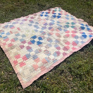 May include: A vintage quilt with a patchwork design in shades of pink, blue, and white. The quilt features a repeating pattern of squares and a pink scalloped edge. The quilt is lying on green grass.