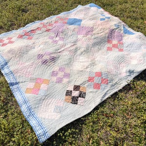 Può includere: Una trapunta vintage con sfondo azzurro e vari quadrati patchwork colorati. La trapunta presenta quadrati rossi, rosa, viola e neri, con un bordo a righe blu e bianco. La trapunta è appoggiata sull'erba verde.