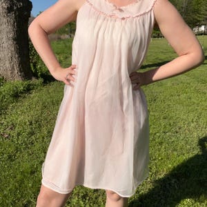 May include: A light pink sleeveless nightgown with a ruffled neckline and small bow accents. The nightgown is made of a sheer, flowing fabric and falls to about knee length. The garment is a vintage style.