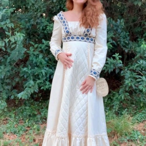 Puede incluir: Vestido largo color crema con mangas largas y escote cuadrado. El vestido presenta detalles bordados en azul y blanco en el escote, los pu&ntilde;os y la cintura. La falda tiene un patr&oacute;n acolchado y un dobladillo con volantes. Una etiqueta est&aacute; adjunta.