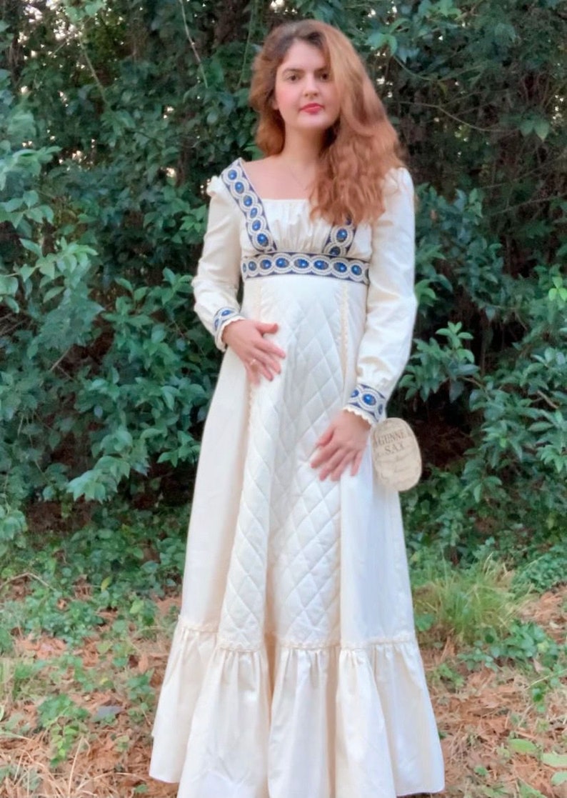 Puede incluir: Vestido largo color crema con mangas largas y escote cuadrado. El vestido presenta detalles bordados en azul y blanco en el escote, los pu&ntilde;os y la cintura. La falda tiene un patr&oacute;n acolchado y un dobladillo con volantes. Una etiqueta est&aacute; adjunta.