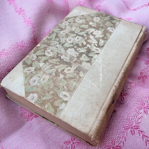 May include: An antique book with a floral patterned cover in shades of green and cream. The spine and edges are a light beige, and the book rests on a pink floral embroidered fabric. The book's cover features a design of white flowers and green leaves.