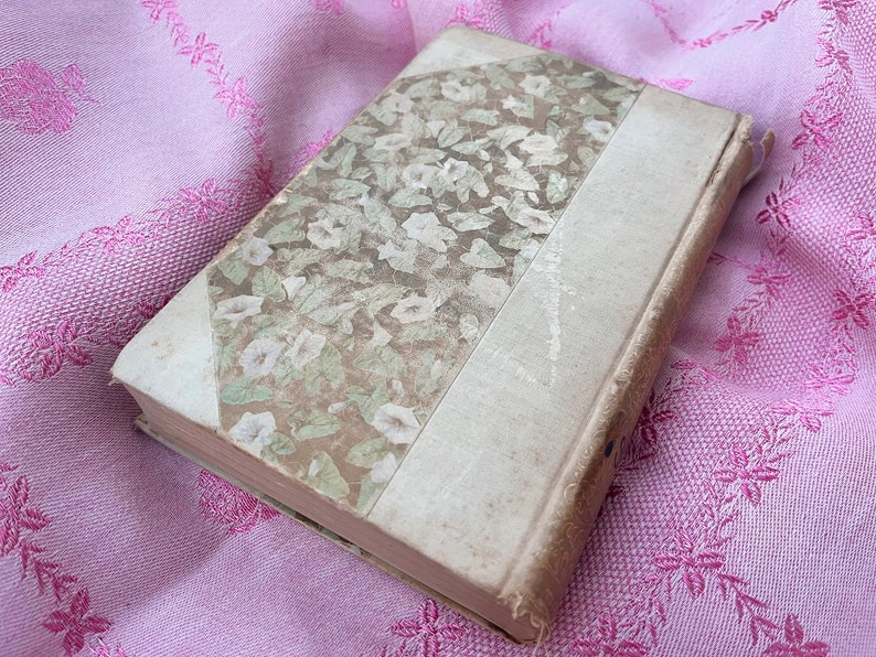 May include: An antique book with a floral patterned cover in shades of green and cream. The spine and edges are a light beige, and the book rests on a pink floral embroidered fabric. The book's cover features a design of white flowers and green leaves.