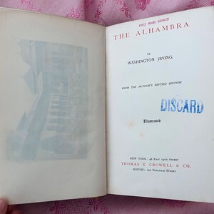 May include: Open book with the title "The Alhambra" by Washington Irving. The cover features a blue and white illustration of a building. The text "DISCARD" is stamped in blue. Published by Thomas Y. Crowell & Co.