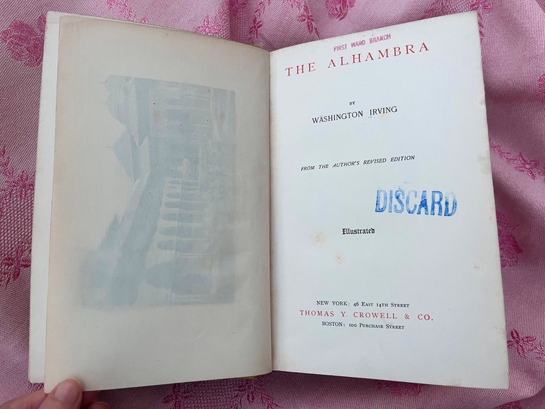 May include: Open book with the title "The Alhambra" by Washington Irving. The cover features a blue and white illustration of a building. The text "DISCARD" is stamped in blue. Published by Thomas Y. Crowell & Co.