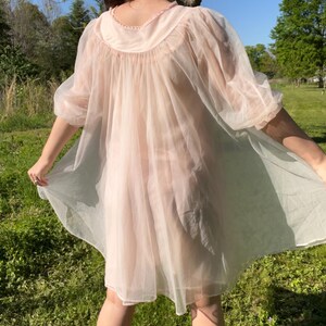May include: A sheer, light pink vintage nightgown with a round neckline and short sleeves. The nightgown is made of a lightweight, flowing fabric. The model is standing in a grassy area with trees in the background.