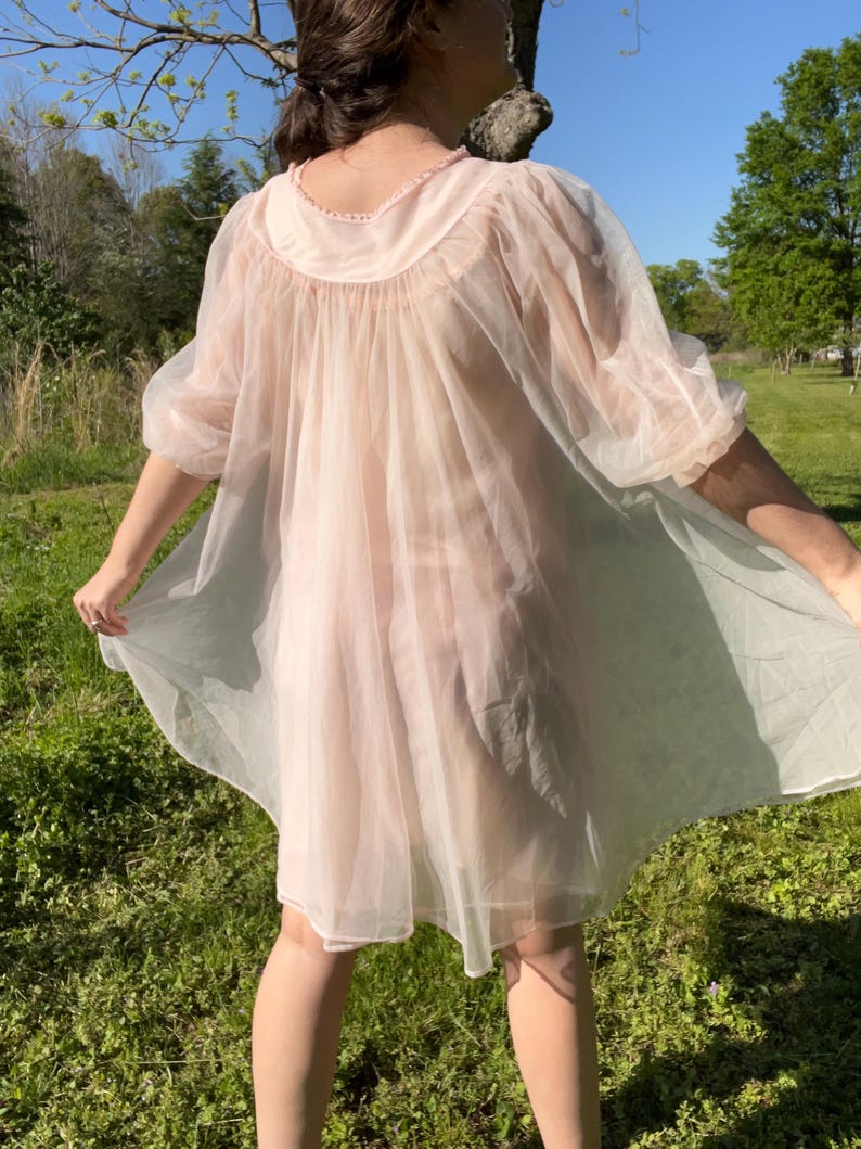 May include: A sheer, light pink vintage nightgown with a round neckline and short sleeves. The nightgown is made of a lightweight, flowing fabric. The model is standing in a grassy area with trees in the background.