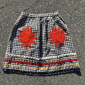 May include: A black and white checkered skirt with two large, orange starburst appliqués. The skirt has a scalloped hemline with white, red, and yellow trim. The skirt is made of a lightweight fabric.