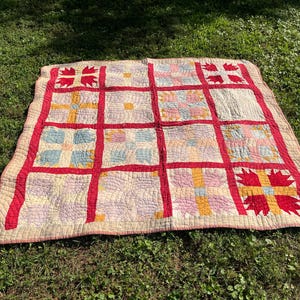 May include: A vintage quilt with a grid pattern, featuring red borders and accents. The quilt's squares contain various pastel colors, including blue, pink, and yellow. The quilt is laid out on a bed of green grass, showcasing its handmade quality.