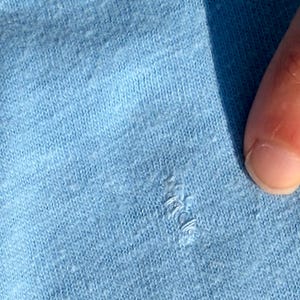 以下が含まれることがあります： 小さな糸のほつれや引きつれがある、ライトブルーのニット生地。素材の質感がはっきりと確認でき、ライトブルーの糸で編まれた織り模様がテキスタイル素材の表面全体にわたって見えています。