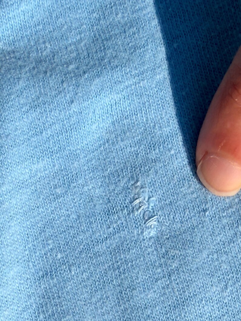以下が含まれることがあります： 小さな糸のほつれや引きつれがある、ライトブルーのニット生地。素材の質感がはっきりと確認でき、ライトブルーの糸で編まれた織り模様がテキスタイル素材の表面全体にわたって見えています。