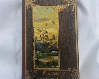 May include: Antique hardcover book titled "Courtship of Miles Standish." The cover features a landscape painting within a gold-trimmed frame, set against a dark wood-grain background. The spine is a dark red color.