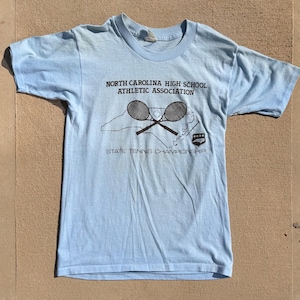May include: A light blue short-sleeved t-shirt featuring a dark graphic print. The design includes the text NORTH CAROLINA HIGH SCHOOL ATHLETIC ASSOCIATION, an outline of the state, two crossed tennis rackets, and STATE TENNIS CHAMPIONSHIP.