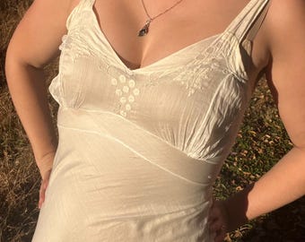 May include: Cream-colored slip dress with spaghetti straps and a V-neckline. The dress features embroidered detailing on the bodice and a fitted waist. A silver necklace with a dark pendant is worn with the dress. The dress appears to be made of a lightweight fabric.