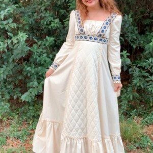 Puede incluir: Vestido largo color crema con mangas largas, escote cuadrado y corpi&ntilde;o acolchado. El vestido presenta detalles bordados en azul y blanco en el escote, la cintura y los pu&ntilde;os, con un dobladillo con volantes. El vestido est&aacute; hecho de una tela suave y fluida.