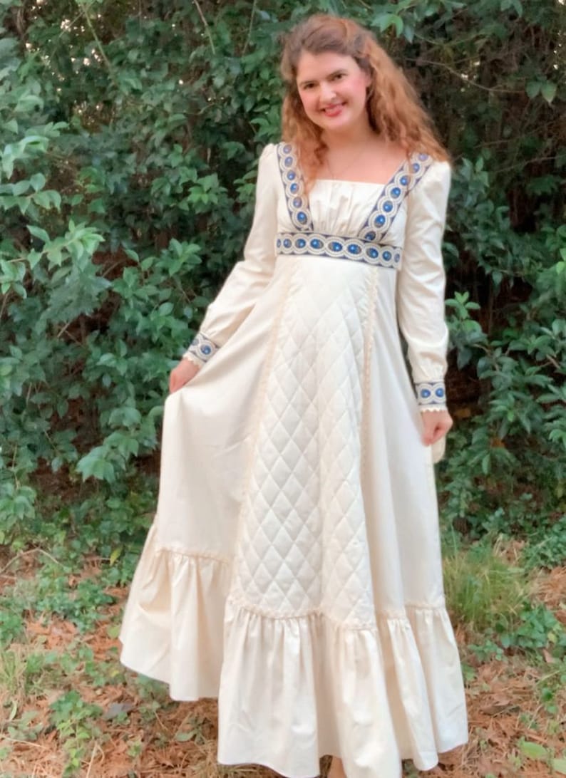 Puede incluir: Vestido largo color crema con mangas largas, escote cuadrado y corpi&ntilde;o acolchado. El vestido presenta detalles bordados en azul y blanco en el escote, la cintura y los pu&ntilde;os, con un dobladillo con volantes. El vestido est&aacute; hecho de una tela suave y fluida.