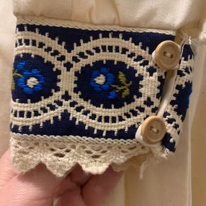 Puede incluir: Primer plano de un pu&ntilde;o de manga color crema con detalles intrincados. El pu&ntilde;o presenta una banda azul marino con patrones florales bordados en azul y verde, bordeada por un ribete de encaje color crema. Dos botones de madera aseguran el pu&ntilde;o.