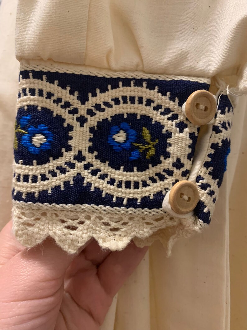 Puede incluir: Primer plano de un pu&ntilde;o de manga color crema con detalles intrincados. El pu&ntilde;o presenta una banda azul marino con patrones florales bordados en azul y verde, bordeada por un ribete de encaje color crema. Dos botones de madera aseguran el pu&ntilde;o.