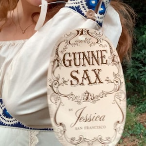 Puede incluir: Vestido color crema con pu&ntilde;os bordados en azul y blanco y una etiqueta que dice "GUNNE SAX by Jessica SAN FRANCISCO". El vestido tiene un ribete de encaje y un escote coraz&oacute;n. Los pu&ntilde;os tienen botones de madera.