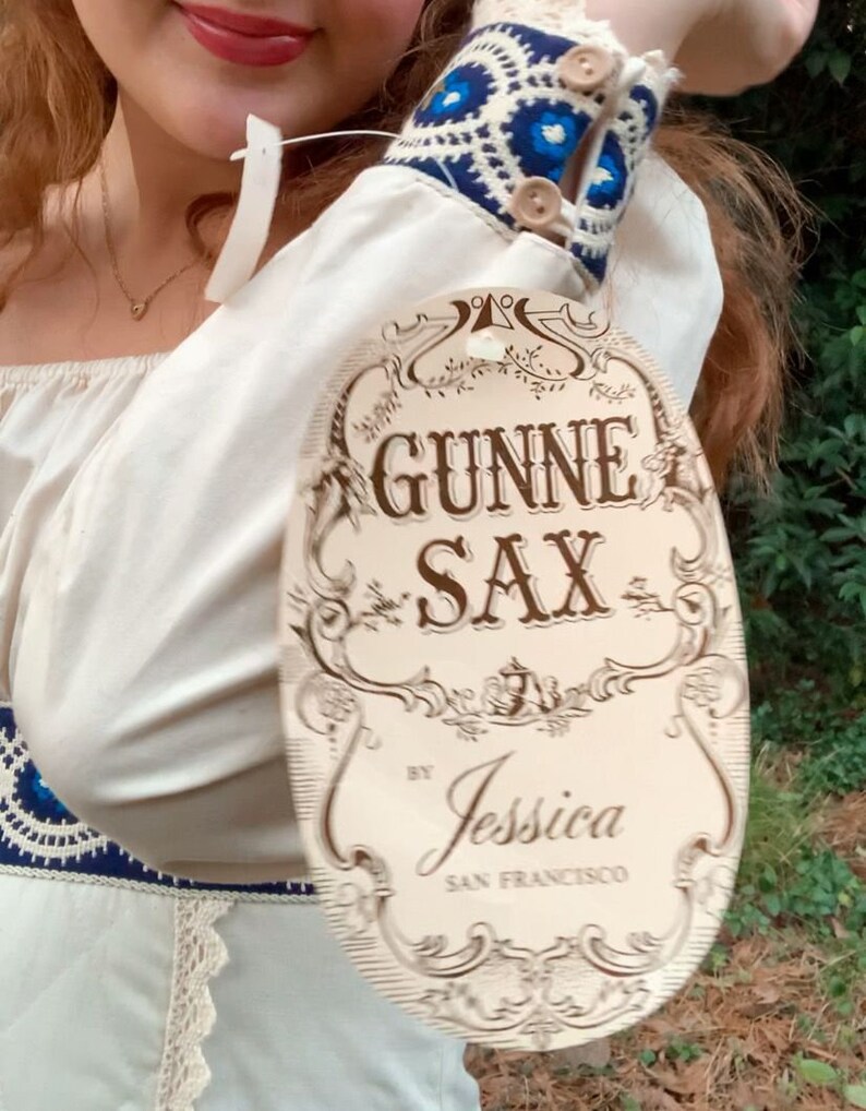 Puede incluir: Vestido color crema con pu&ntilde;os bordados en azul y blanco y una etiqueta que dice "GUNNE SAX by Jessica SAN FRANCISCO". El vestido tiene un ribete de encaje y un escote coraz&oacute;n. Los pu&ntilde;os tienen botones de madera.