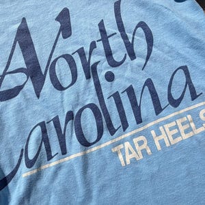 以下が含まれることがあります： 大きなスタイリッシュなフォントで濃い青色の「North Carolina」という文字がプリントされた水色のTシャツ。その下には白い線で下線が引かれた白い「TAR HEELS」という文字があり、小さな登録商標マークも含まれています。