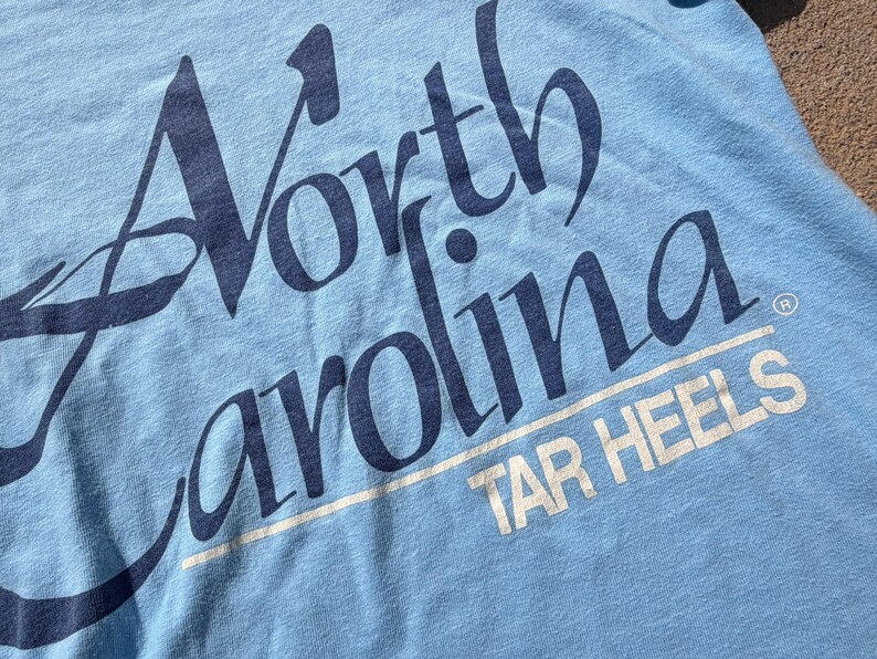 以下が含まれることがあります： 大きなスタイリッシュなフォントで濃い青色の「North Carolina」という文字がプリントされた水色のTシャツ。その下には白い線で下線が引かれた白い「TAR HEELS」という文字があり、小さな登録商標マークも含まれています。