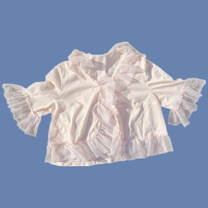 May include: A light pink, open-front jacket with ruffled trim and bell sleeves. The jacket features a V-neckline and a delicate, sheer ruffle detail along the front and sleeve edges. The fabric appears soft and lightweight, suitable for layering.