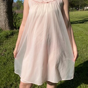 May include: A vintage-style, pale pink nightgown. The sleeveless garment features a ruffled neckline with a small bow and a flowing, sheer skirt. The nightgown is made of a lightweight, semi-transparent fabric.