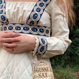 Puede incluir: Vestido color crema con bordado floral azul en el escote, la cintura y los pu&ntilde;os. El vestido presenta un escote cuadrado, mangas largas y un corpi&ntilde;o acolchado. Una etiqueta dice "GUNNE SAX by Jessica San Francisco."