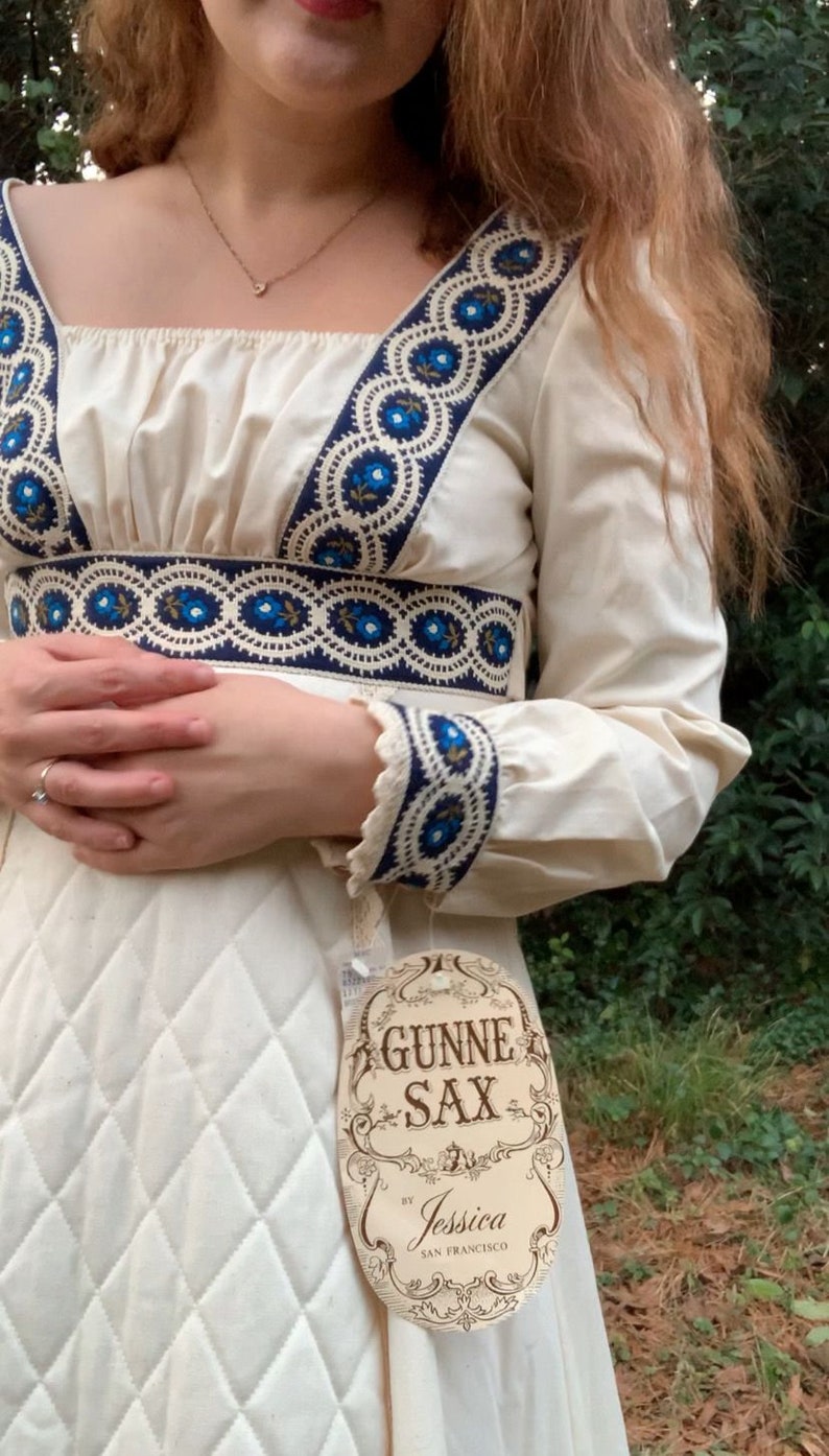 Puede incluir: Vestido color crema con bordado floral azul en el escote, la cintura y los pu&ntilde;os. El vestido presenta un escote cuadrado, mangas largas y un corpi&ntilde;o acolchado. Una etiqueta dice "GUNNE SAX by Jessica San Francisco."