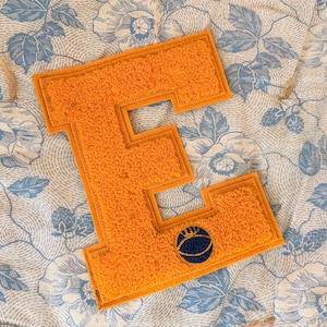 May include: An orange letter "E" patch with a navy blue basketball emblem. The patch has a textured, fuzzy appearance and a contrasting orange border. The background features a floral pattern in blue and white.