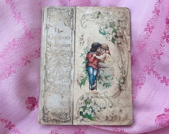 May include: Antique book titled "The Children's Shakespeare" with a faded cover. The cover features an illustration of two children embracing, surrounded by floral elements. The book is set against a pink floral patterned fabric.
