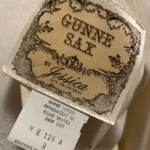 Puede incluir: Primer plano de una etiqueta de ropa vintage. La etiqueta dice "GUNNE SAX by Jessica, San Francisco". Texto adicional incluye instrucciones de lavado e informaci&oacute;n de tallas. La etiqueta es de color crema con detalles marrones ornamentados.