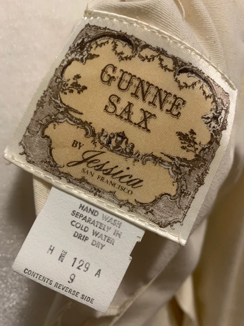 Puede incluir: Primer plano de una etiqueta de ropa vintage. La etiqueta dice "GUNNE SAX by Jessica, San Francisco". Texto adicional incluye instrucciones de lavado e informaci&oacute;n de tallas. La etiqueta es de color crema con detalles marrones ornamentados.