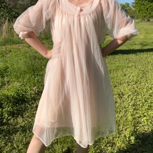 May include: A vintage, sheer, pale pink nightgown. The nightgown features a round neckline with delicate lace trim and a small button closure. The sleeves are short and puffy, and the skirt falls to the mid-calf. The fabric appears lightweight and flowing.