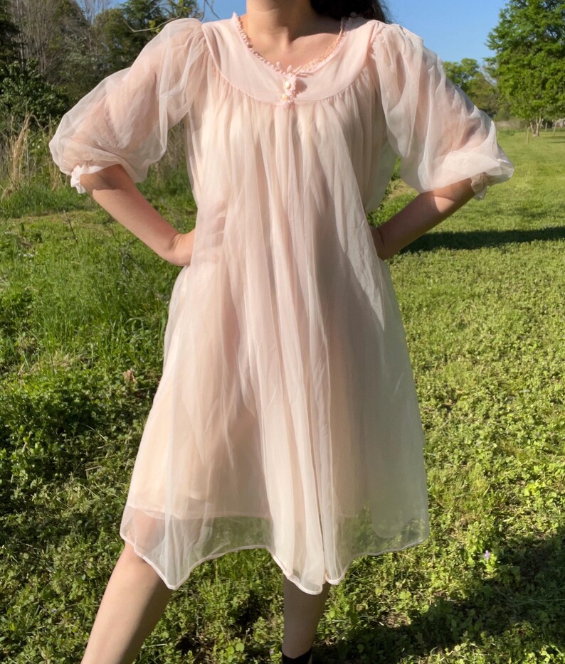 May include: A vintage, sheer, pale pink nightgown. The nightgown features a round neckline with delicate lace trim and a small button closure. The sleeves are short and puffy, and the skirt falls to the mid-calf. The fabric appears lightweight and flowing.