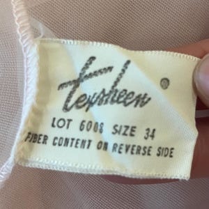 May include: A close-up of a tag with the word "Texsheen" in cursive. The tag also includes the text "LOT 6008 SIZE 34 FIBER CONTENT ON REVERSE SIDE". The tag is on a pink fabric.