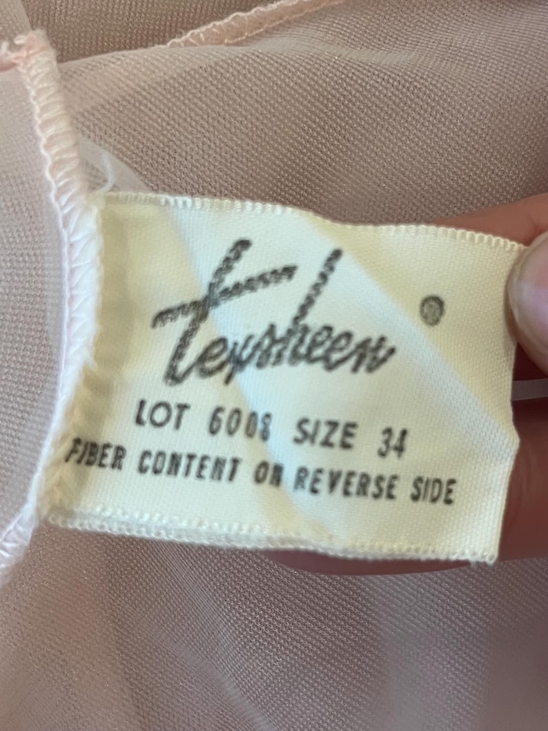 May include: A close-up of a tag with the word "Texsheen" in cursive. The tag also includes the text "LOT 6008 SIZE 34 FIBER CONTENT ON REVERSE SIDE". The tag is on a pink fabric.
