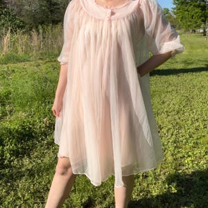 May include: A light pink vintage nightgown with a sheer, flowing design. The nightgown features a round neckline with delicate trim and short, puffed sleeves. The fabric appears lightweight and airy, perfect for a comfortable night's sleep. The garment is knee-length.