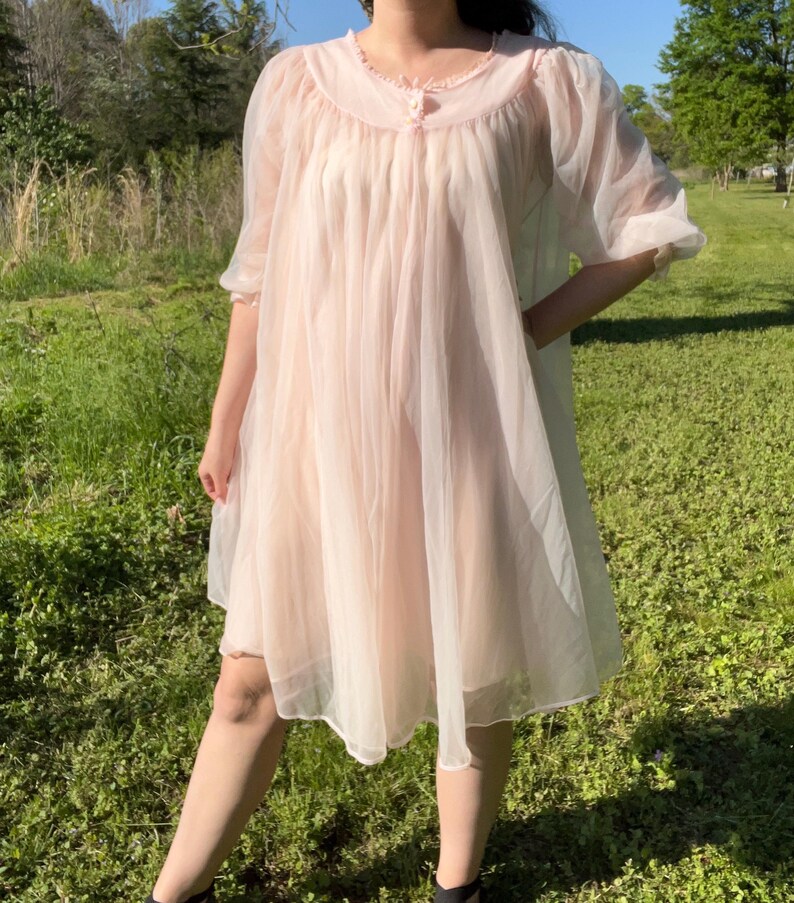 May include: A light pink vintage nightgown with a sheer, flowing design. The nightgown features a round neckline with delicate trim and short, puffed sleeves. The fabric appears lightweight and airy, perfect for a comfortable night's sleep. The garment is knee-length.